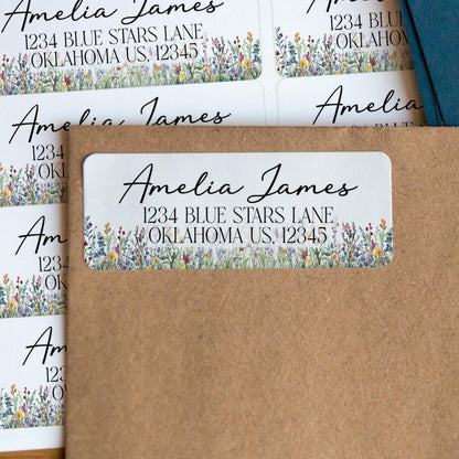 May include: White address labels with the name "Amelia James" in elegant script. The labels include an address and a floral border with blue, yellow, and purple flowers. The labels are on a brown envelope.