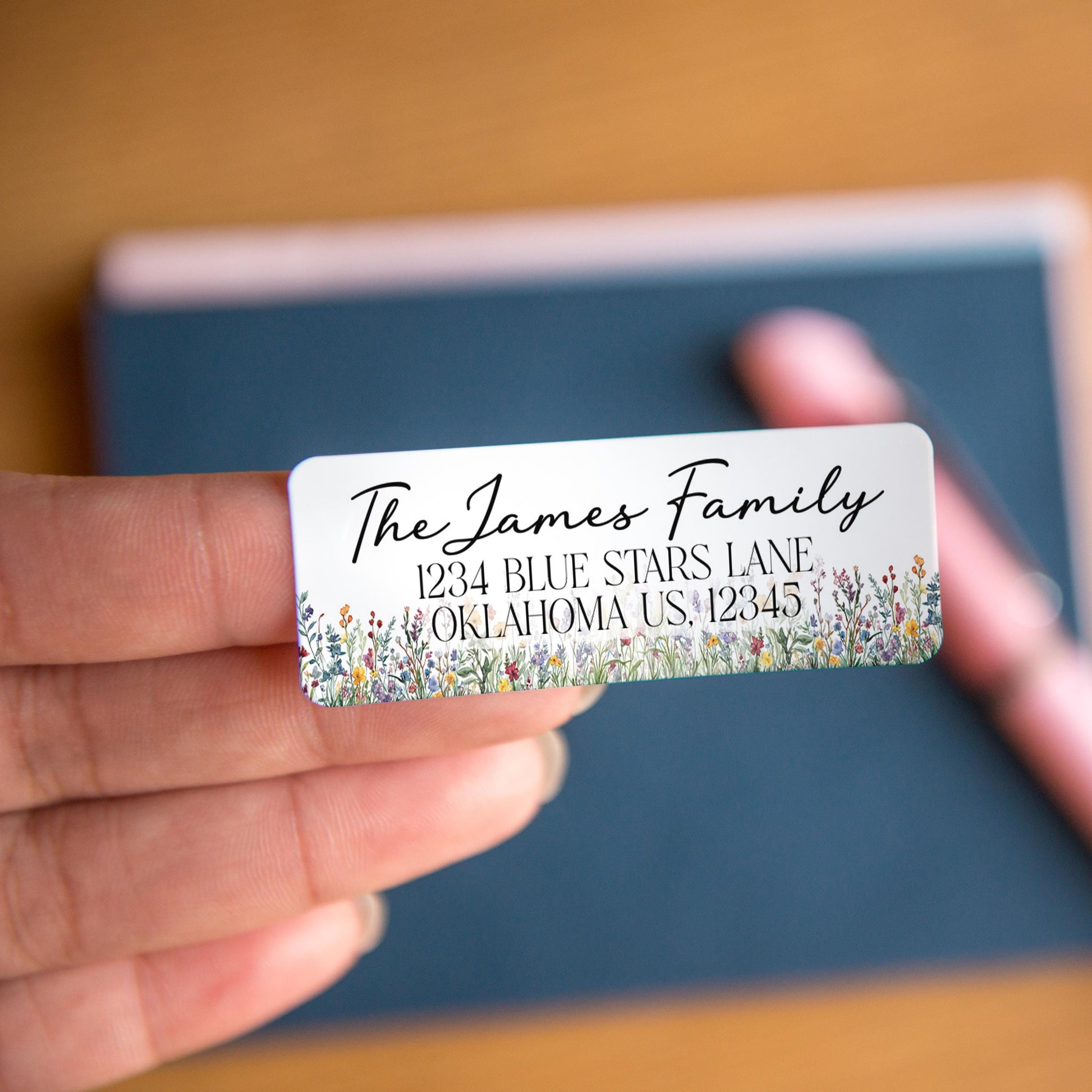 May include: A rectangular white address label with the text "The James Family" in elegant script. The label includes the address "1234 BLUE STARS LANE, OKLAHOMA US, 12345" in a simple font. The bottom edge is decorated with a floral border.
