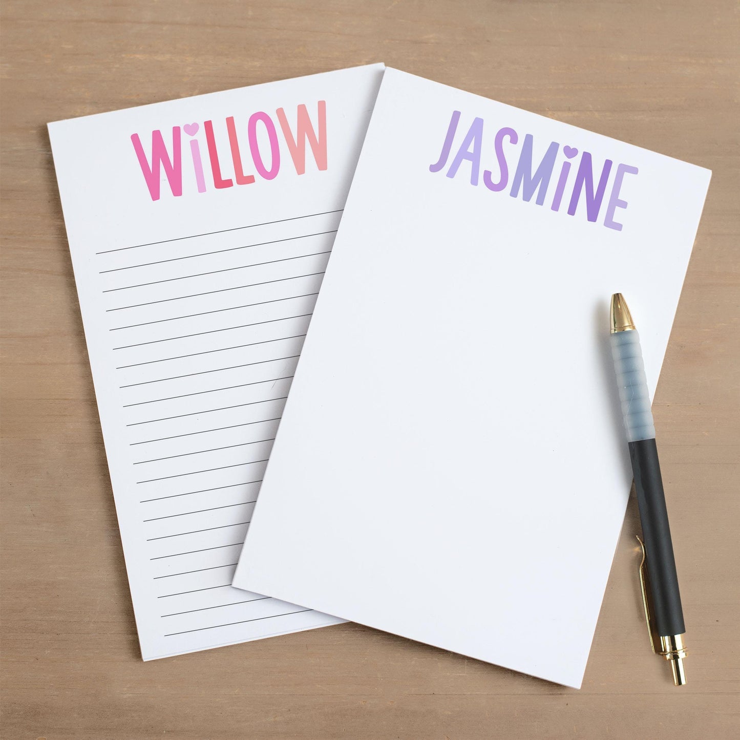 May include: Two white notepad sets with lined pages. The top of each notepad has a colorful gradient design with a heart and a name. The notepad on the left has the name 'WILLOW' in pink and red. The notepad on the right has the name 'JASMINE' in purple and blue. A black pen with a gold tip is lying on the right notepad.