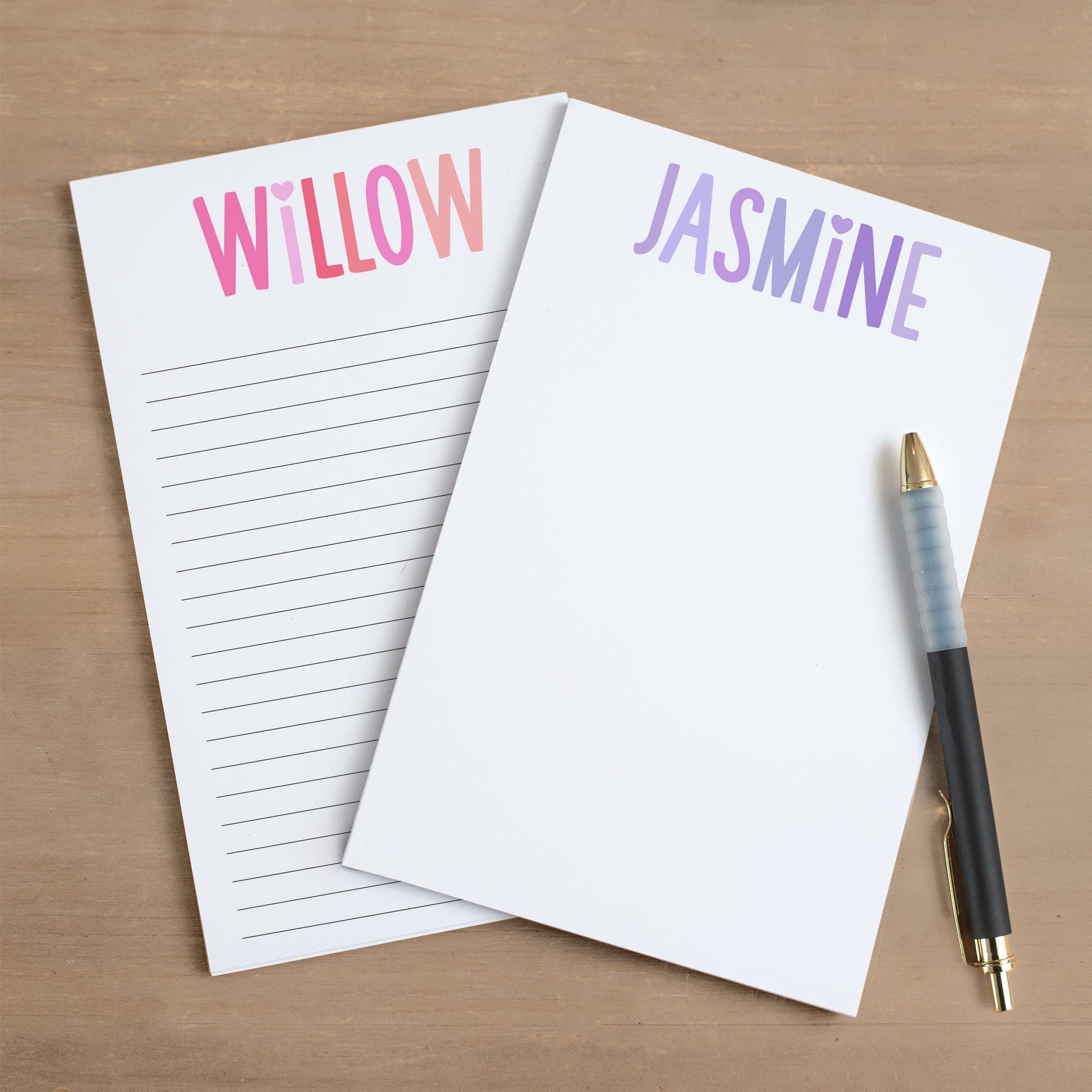 May include: Two white notepad sets with lined pages. The top of each notepad has a colorful gradient design with a heart and a name. The notepad on the left has the name 'WILLOW' in pink and red. The notepad on the right has the name 'JASMINE' in purple and blue. A black pen with a gold tip is lying on the right notepad.