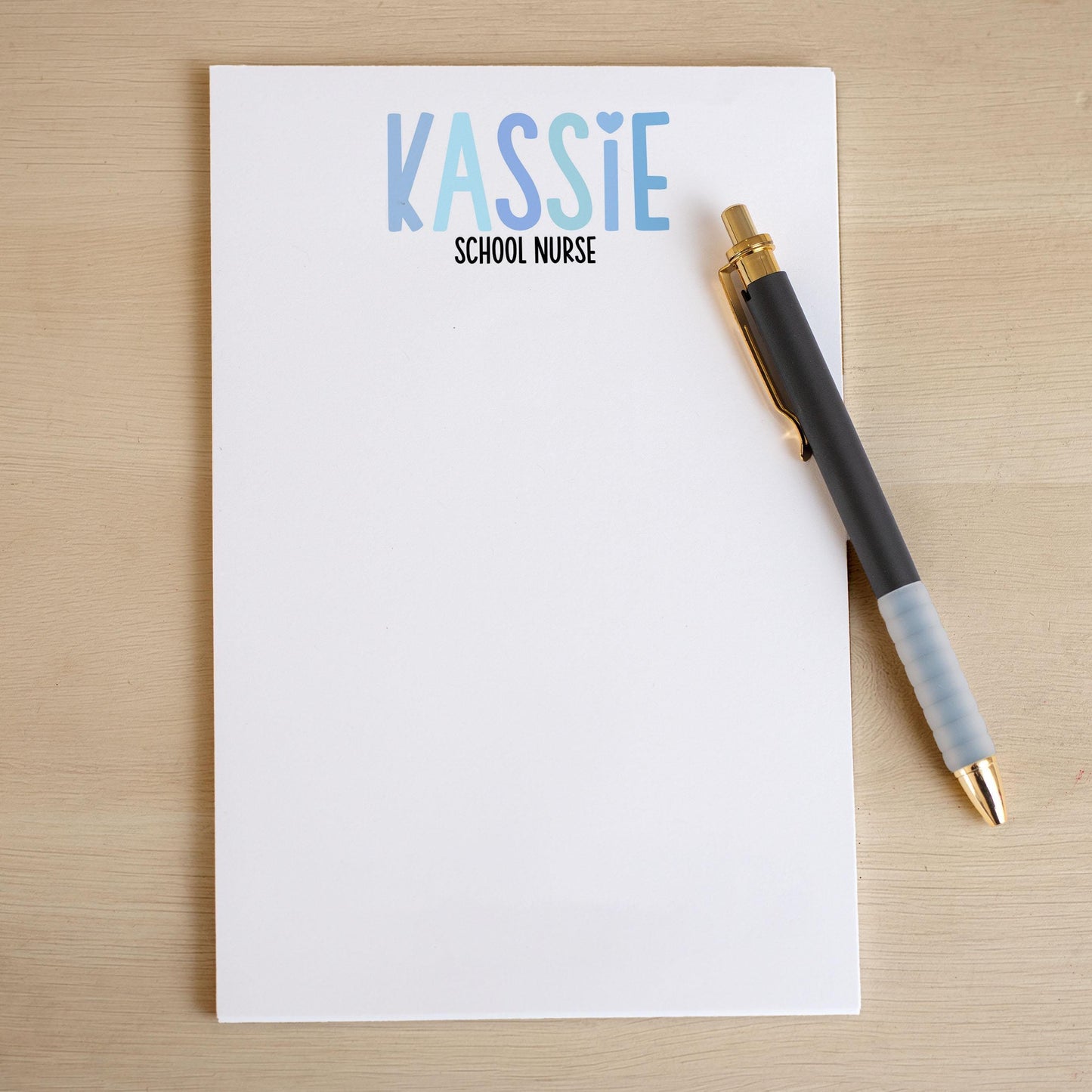 May include: A white notepad with the text 'KASSIE SCHOOL NURSE' printed in blue and light blue lettering. A black pen with a gold top is lying on the notepad.